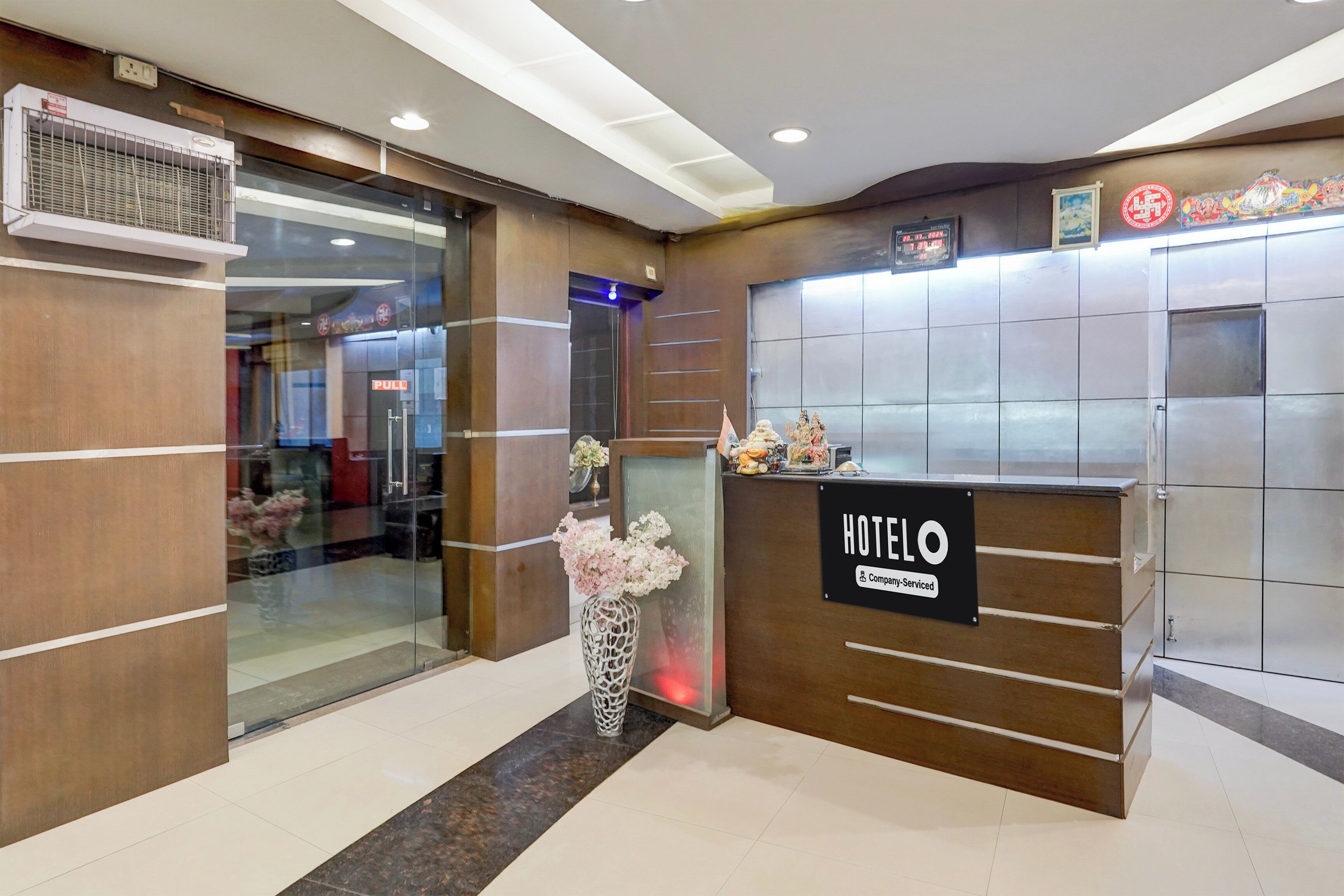 Un mostrador de recepción de hotel con un cartel que indica servicio de empresa. Hay una entrada por puerta de cristal, un área de recepción con un jarrón de flores y un reloj digital que muestra la hora.