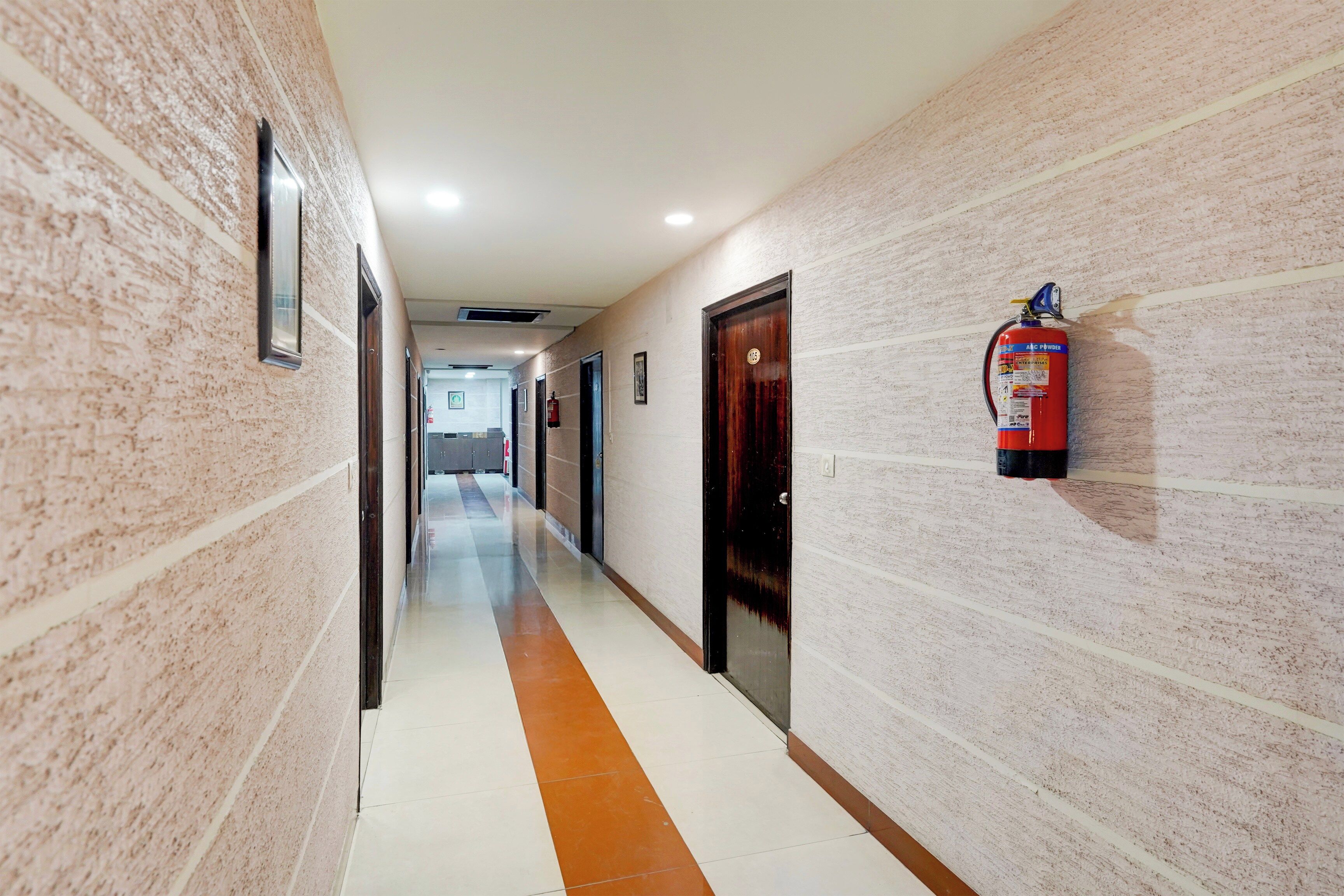 Un pasillo con un extintor montado en la pared, una puerta con placa de número y suelo de baldosas.