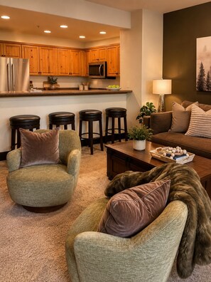 A modern living room with a green armchair, a brown sofa, and a coffee table. There are four bar stools, a lamp, and a painting on the wall. - Bear Creek Lodge 303a 1 Bedroom Condo (Telluride)