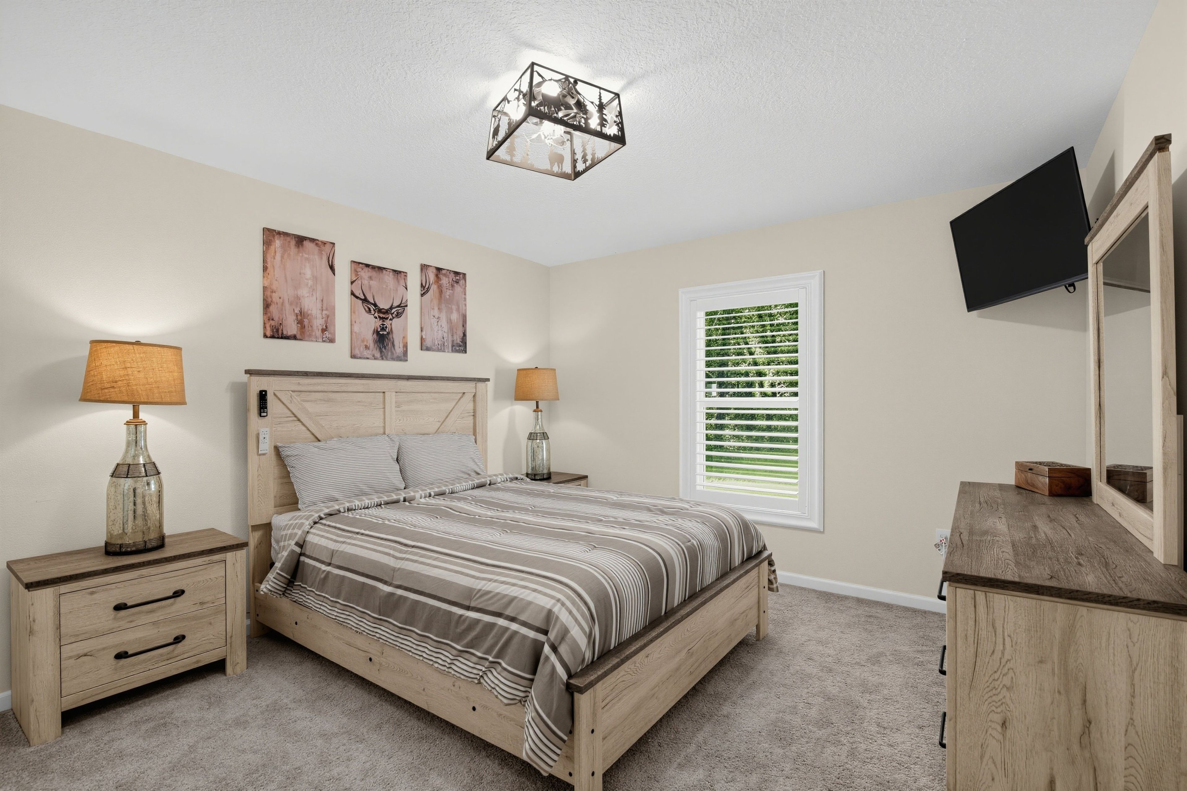 A bedroom with a bed, two bedside tables, a lamp, a TV, a window with blinds, and artwork on the wall.