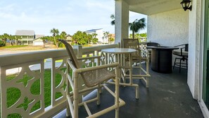 A covered outdoor seating area with chairs and a table, overlooking a street with buildings and palm trees. - 525 El Matador 1 Bedroom Condo (Fort Walton Beach)