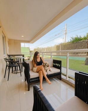 A woman sitting on a sofa with a dog, a dining table, chairs, and a view of a fence and greenery. - VG Fun Praia do Futuro (Fortaleza)