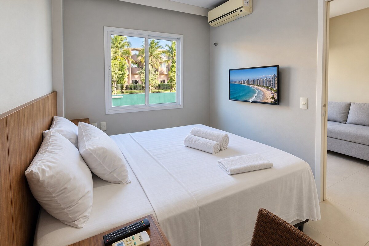 A hotel room with a bed, a TV, a remote control, and a window with a view of a beach and palm trees.