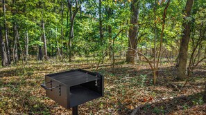 A portable grill in a forested area with trees and leaf-covered ground. - Sleeping Bear Cottage-a brand new getaway (Mentone)