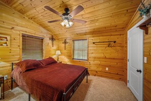 Quarto de cabana de madeira com uma cama, duas janelas com persianas, um ventilador de teto e uma porta branca.