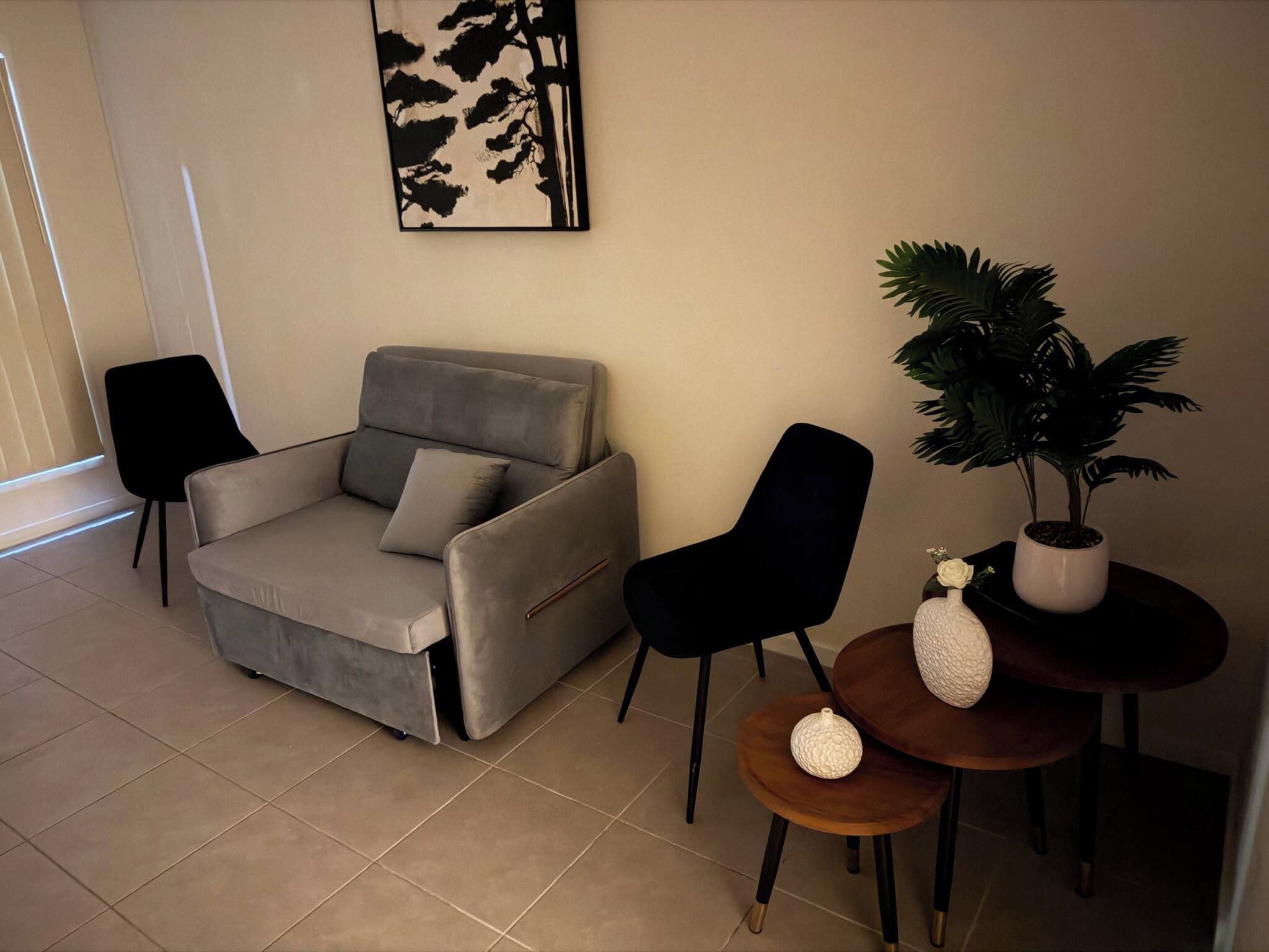A living room with a grey sofa, two black chairs, a small round table with a potted plant, and a wall-mounted abstract art piece.