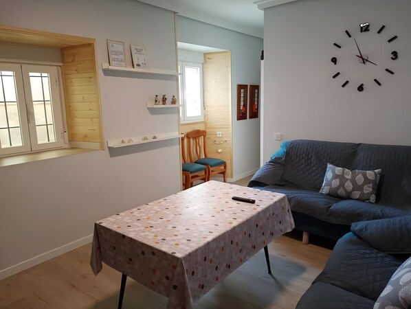 A living room with a sofa, a table with a patterned tablecloth, a clock on the wall, and a window with blinds. - Apartment 'Alojamiento Rural La Tahona Gorafe' with Wi-Fi (Gorafe)
