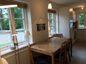 Ein Essbereich mit einem Holztisch und Stühlen, ein Fenster mit Blick auf Grünflächen, eine Wanduhr und eine Küche mit Schränken und Geräten.