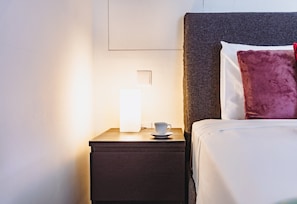 A neatly made bed with a dark headboard, a white pillow, and a red accent pillow. A bedside table with a cup and saucer is illuminated by a lamp. - Mercury Suites Prestige Stay, Pool Access (St. Julian's)