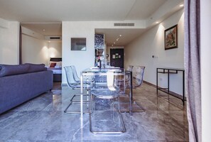 Una sala moderna con un sofá, un comedor con una mesa y sillas de vidrio, y una cocina al fondo.