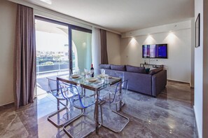 A modern living space with a glass dining table, clear chairs, a grey sofa, and a flat-screen TV mounted on the wall. - Mercury Suites 2BR Luxury Hideaway, Pool Access (St. Julian's)