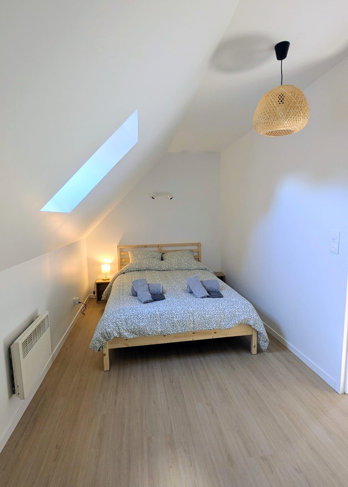 A bedroom with a sloped ceiling, a bed with a patterned bedspread, a bedside lamp, and a woven light fixture.