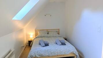 A bedroom with a sloped ceiling, a bed with a patterned bedspread, a bedside lamp, and a woven light fixture.