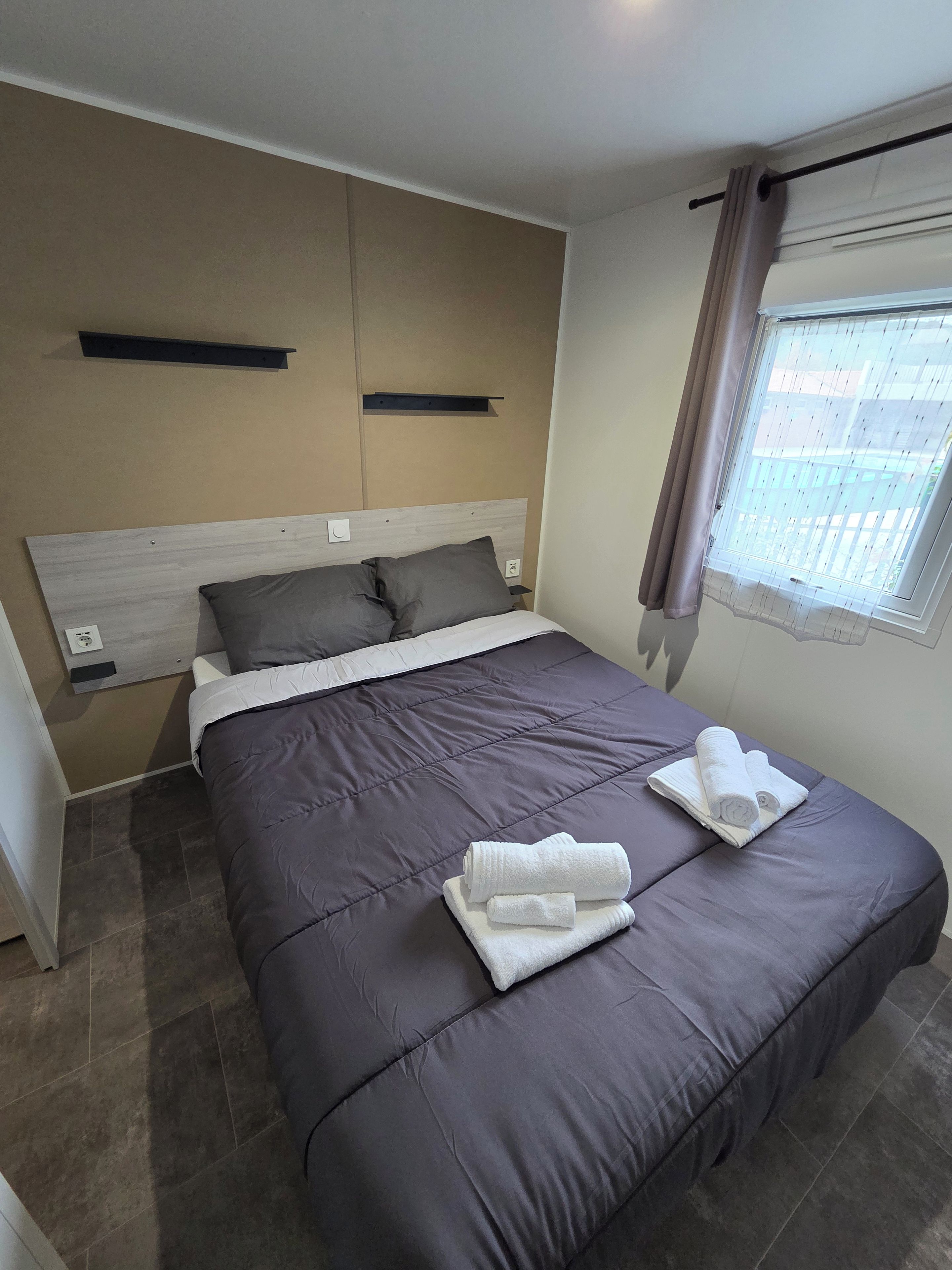 A neatly made bed with a grey duvet, two pillows, and two rolled towels on the bed.