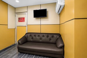 Un área de espera con un sofá de cuero marrón, una puerta cerrada, un televisor montado y una unidad de aire acondicionado en la pared.