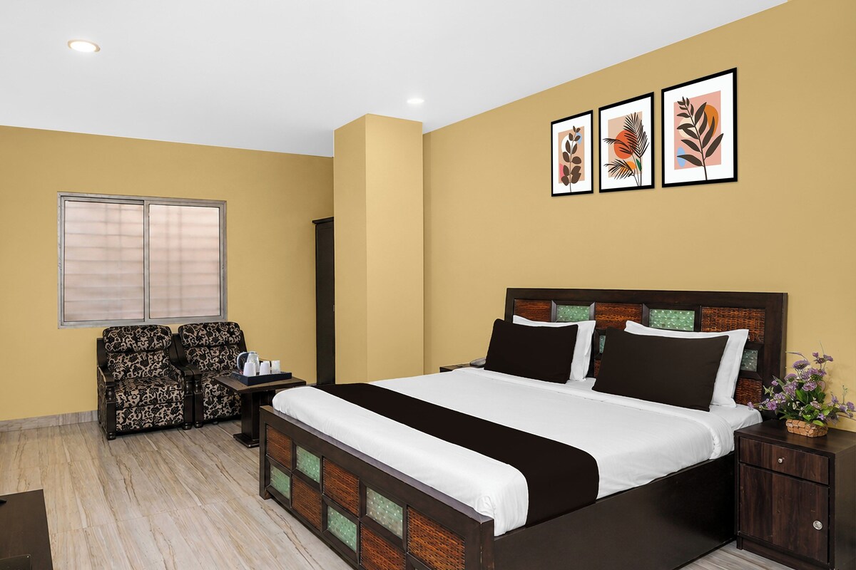 Una habitación de hotel con cama, mesitas de noche, un rincón de estar con sillones estampados, una ventana con persianas y decoración en la pared.