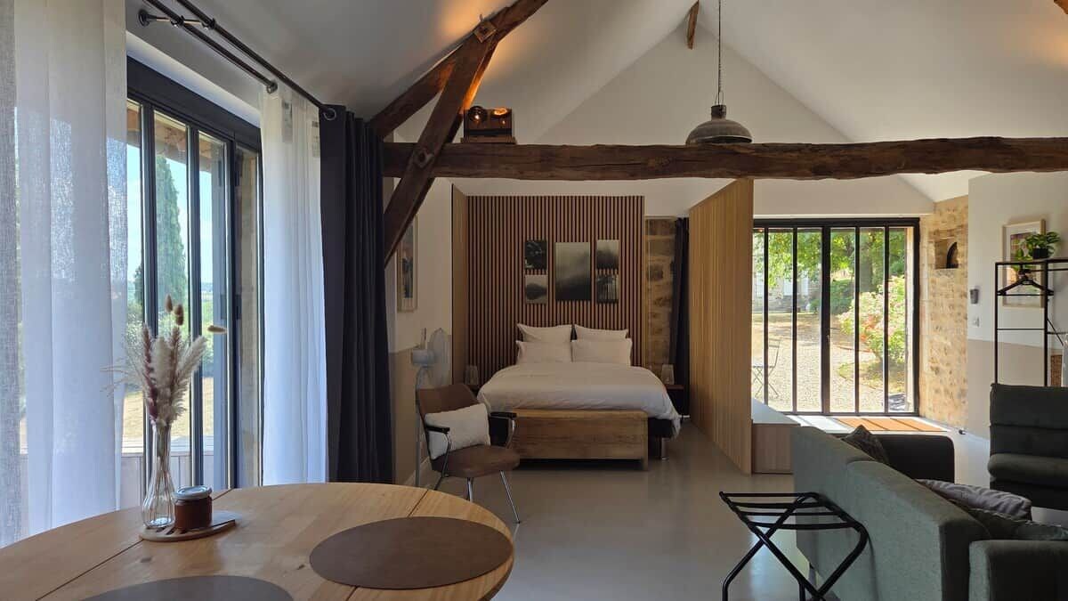 A bedroom with a bed, a wooden table, a chair, and a sofa. There is a window with curtains and a view of the outdoors.
