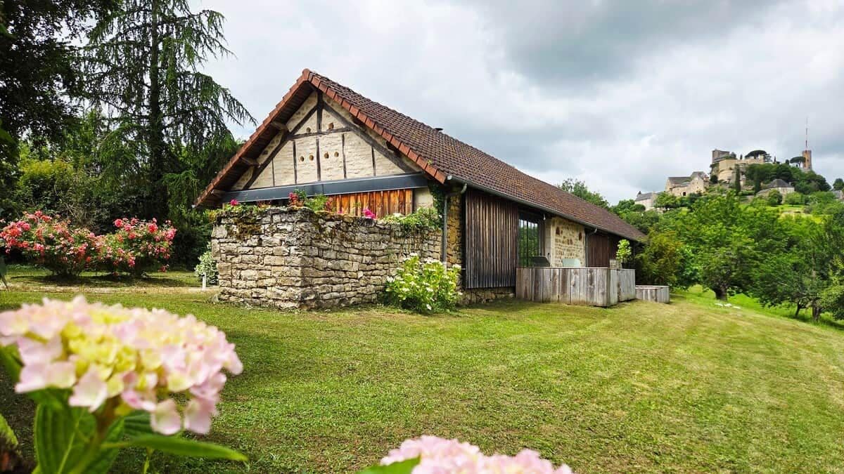 Ein Steinhaus mit geneigtem Dach, umgeben von einem gepflegten Rasen und blühenden Pflanzen.