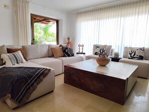 A modern living room with a sectional sofa, a coffee table, and a large window with curtains. - Esta amplia villa de 3 habitaciones ofrece espectaculares vistas al océano, una alberca privada tipo plunge, acceso directo a la playa y acceso a las increíbles amenidades del resort, incluyendo 8 albercas, spa,  restaurante y mucho mas (Cruz de Huanacaxtle)