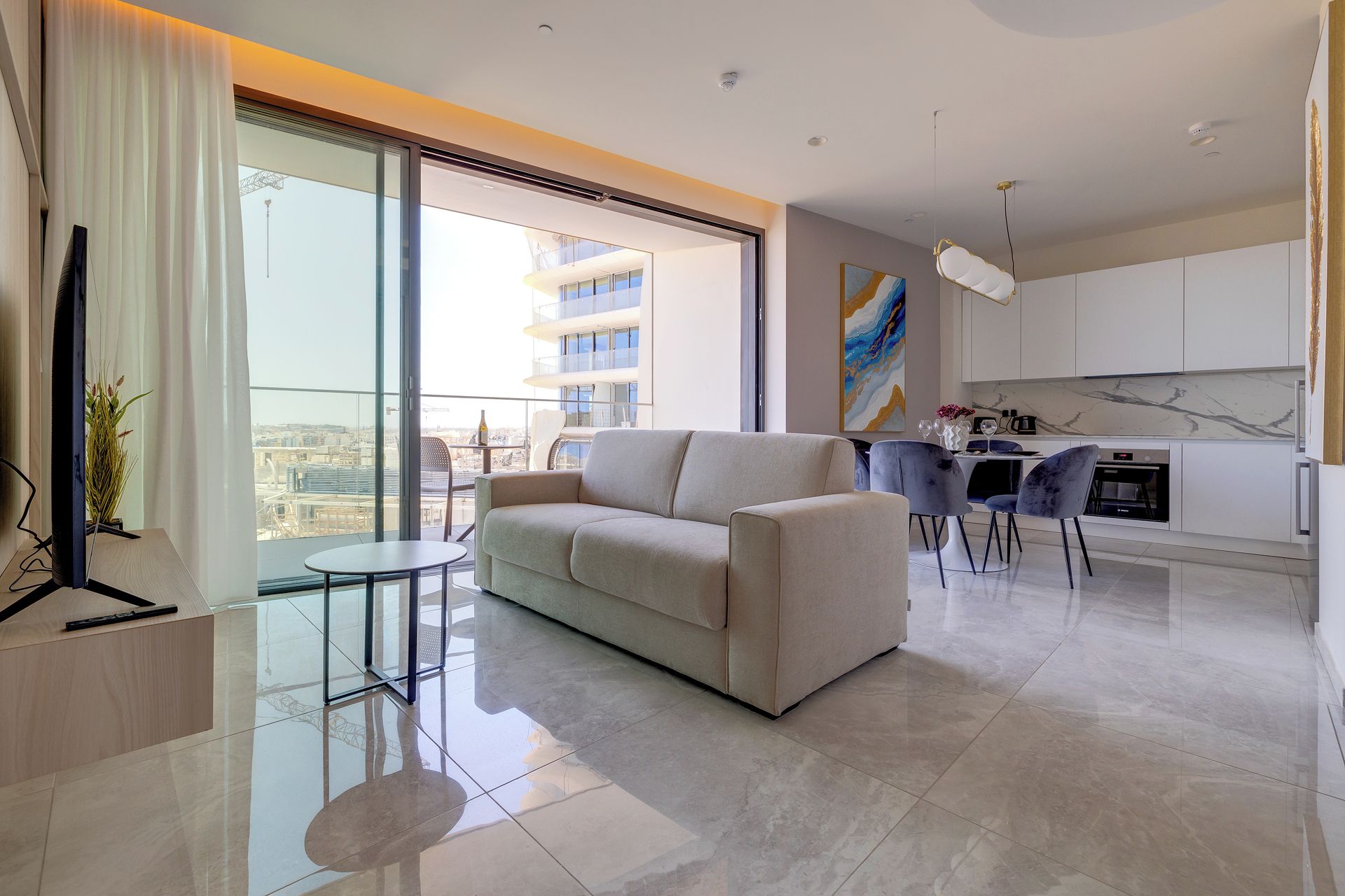 A modern living room with a sofa, a small table, and a TV. There's a balcony with a view of buildings.