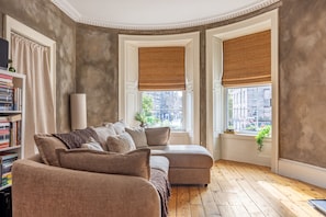 A room with a large sectional sofa, a bookshelf, and two windows with brown blinds. - Lovely Flat near Royal Yacht Britannia (Edinburgh)