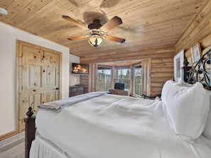 Ein Schlafzimmer einer Holzhütte mit Bett, Deckenventilator, Fernseher und Blick ins Freie.