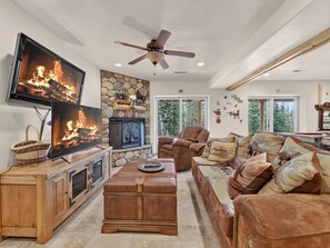 A living room with a fireplace, a television, a ceiling fan, and a stone wall. - Chair 5 Chalet | True Ski In/ Ski Out + Hot Tub (Brian Head)