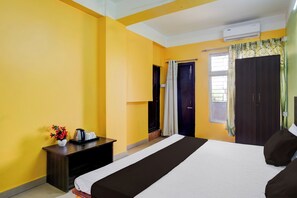 Habitación de hotel con cama, mesita de noche con tetera, una planta pequeña y ventana con cortinas.