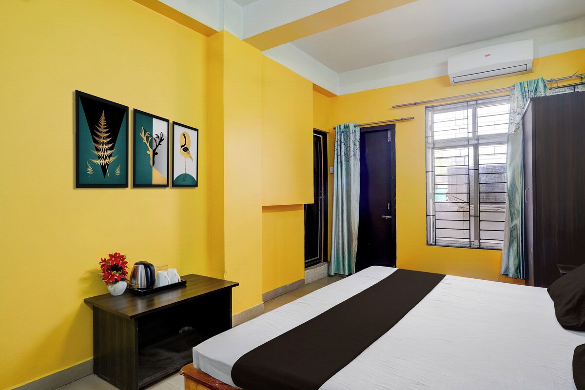 A hotel room with a bed, a bedside table with a kettle, cups, and a vase of flowers. There are three framed artworks on the wall, a window with curtains, and an air conditioning unit.