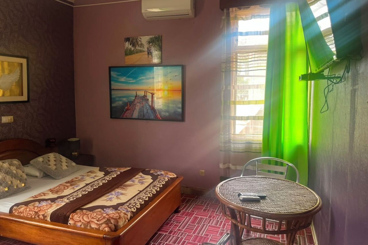 Ein Schlafzimmer mit Bett, einem Bild, einem Fenster mit Vorhängen und einem kleinen Tisch.