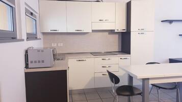 Et kompakt køkken med hvide skabe, en lille bordplade og et spiseområde med et bord og stole.