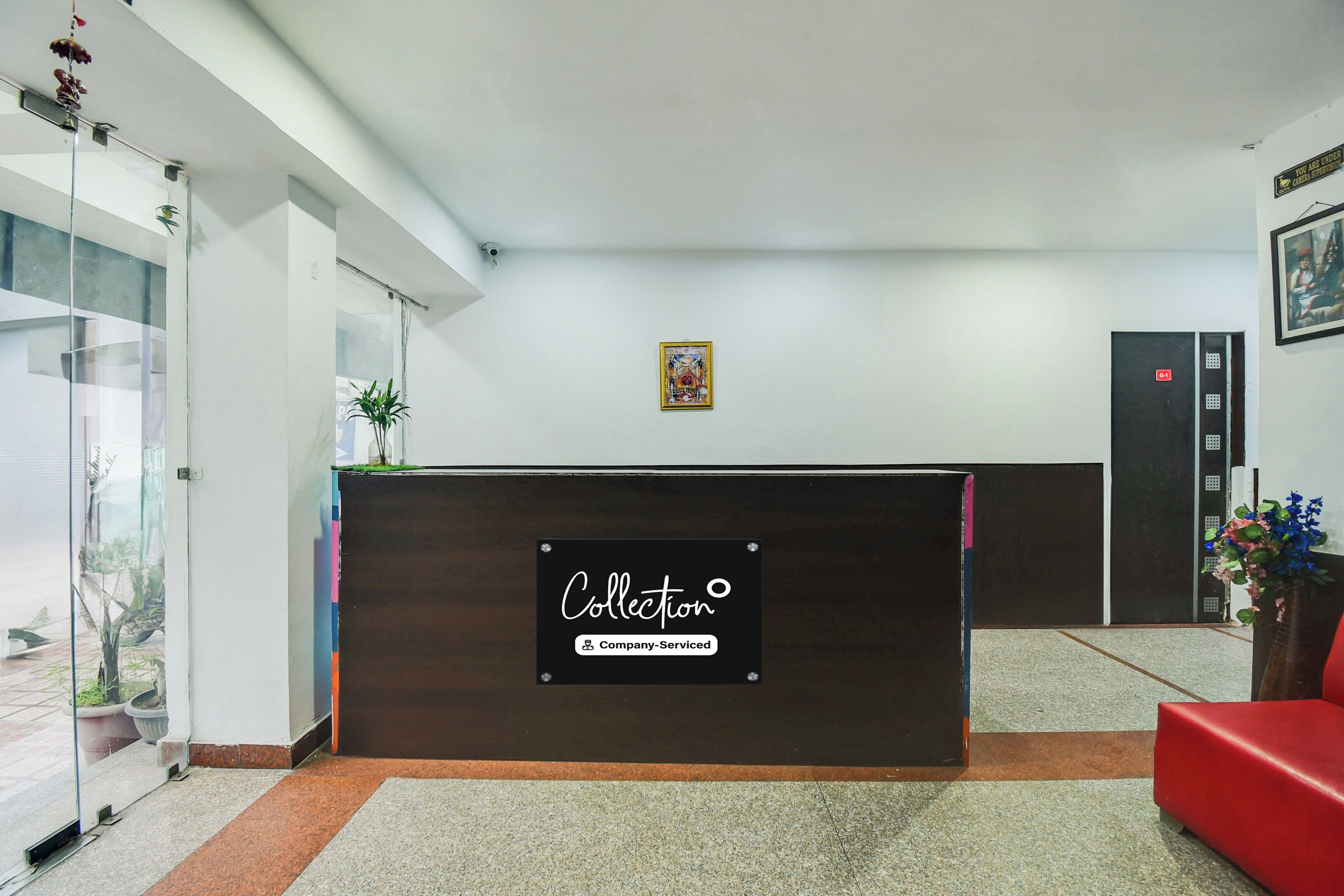 A reception desk with a sign indicating it is company-serviced. There is a framed picture on the wall, a potted plant, and a red chair.