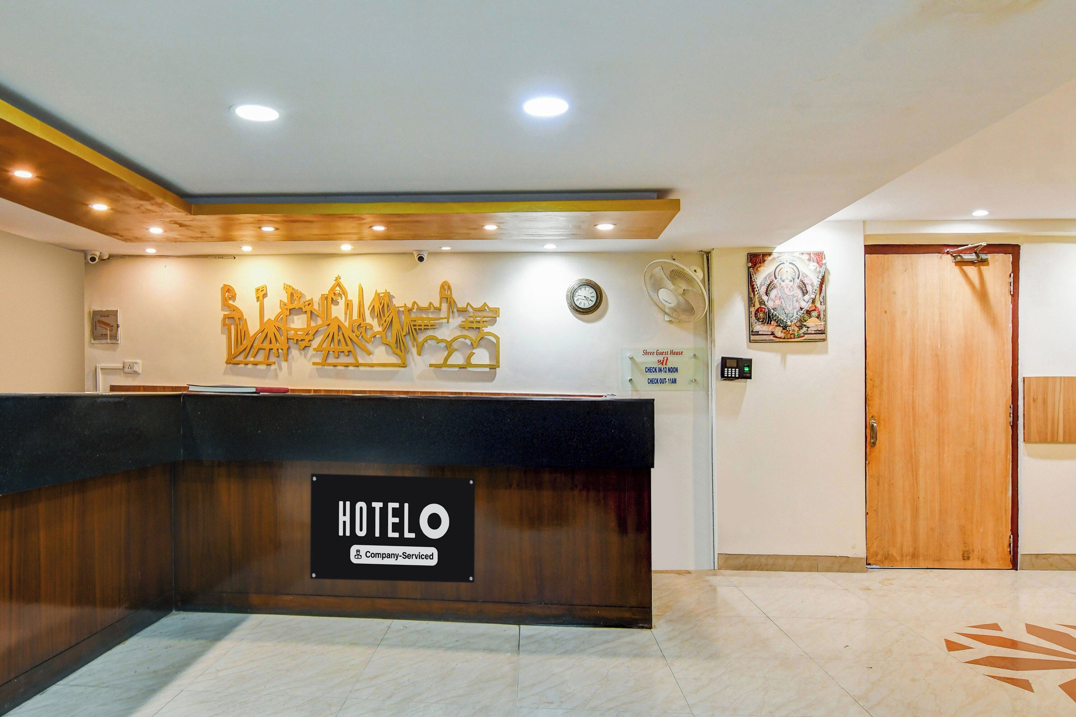 A hotel reception desk with a sign indicating it is company-serviced. There is a clock and a sign for 'Shree Guest House' with check-in and check-out times.