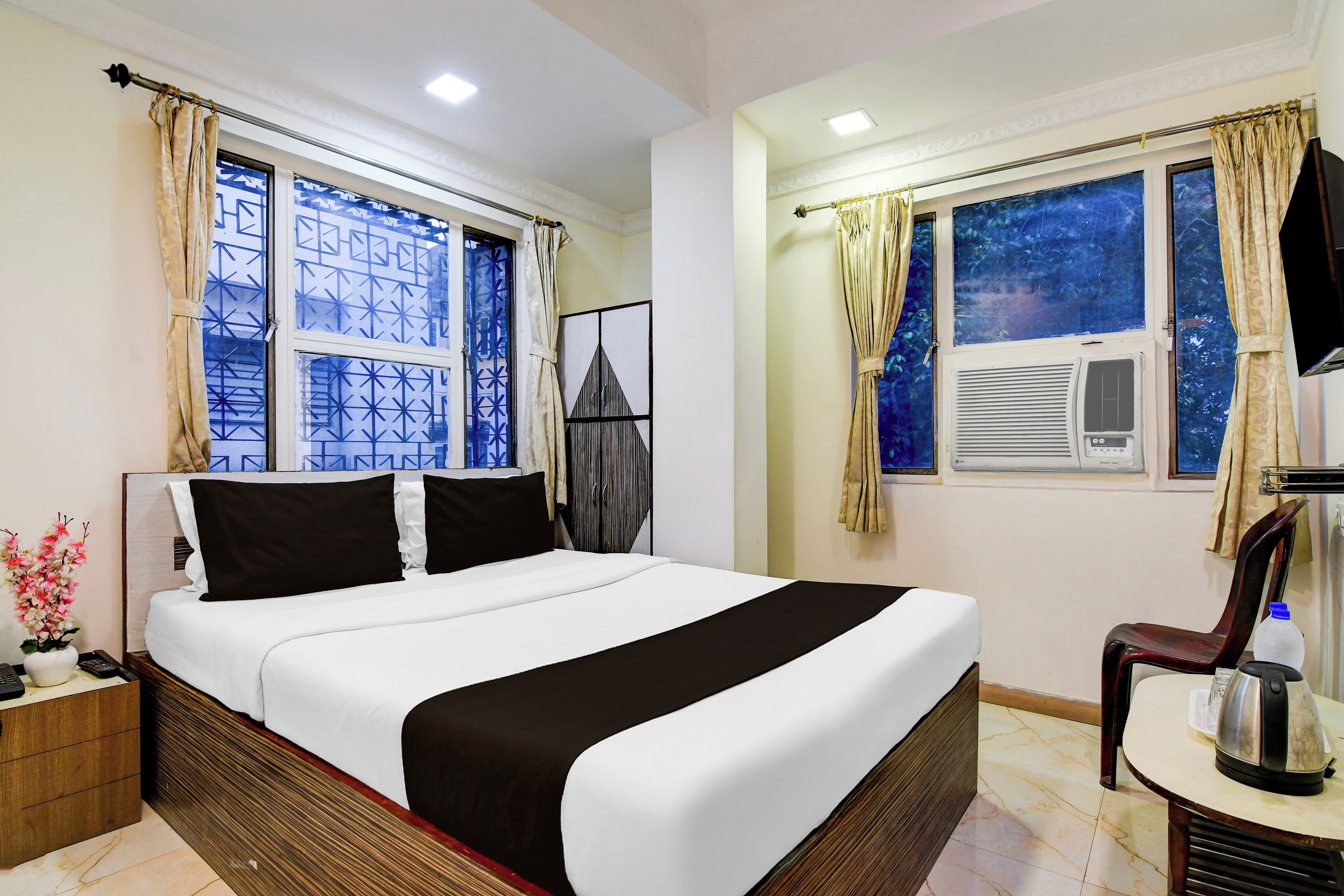 A hotel room with a bed, a desk with a kettle, a chair, a TV, and two windows with curtains.