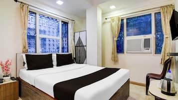 Habitación de hotel con cama, escritorio con tetera, silla, televisor y dos ventanas con cortinas.