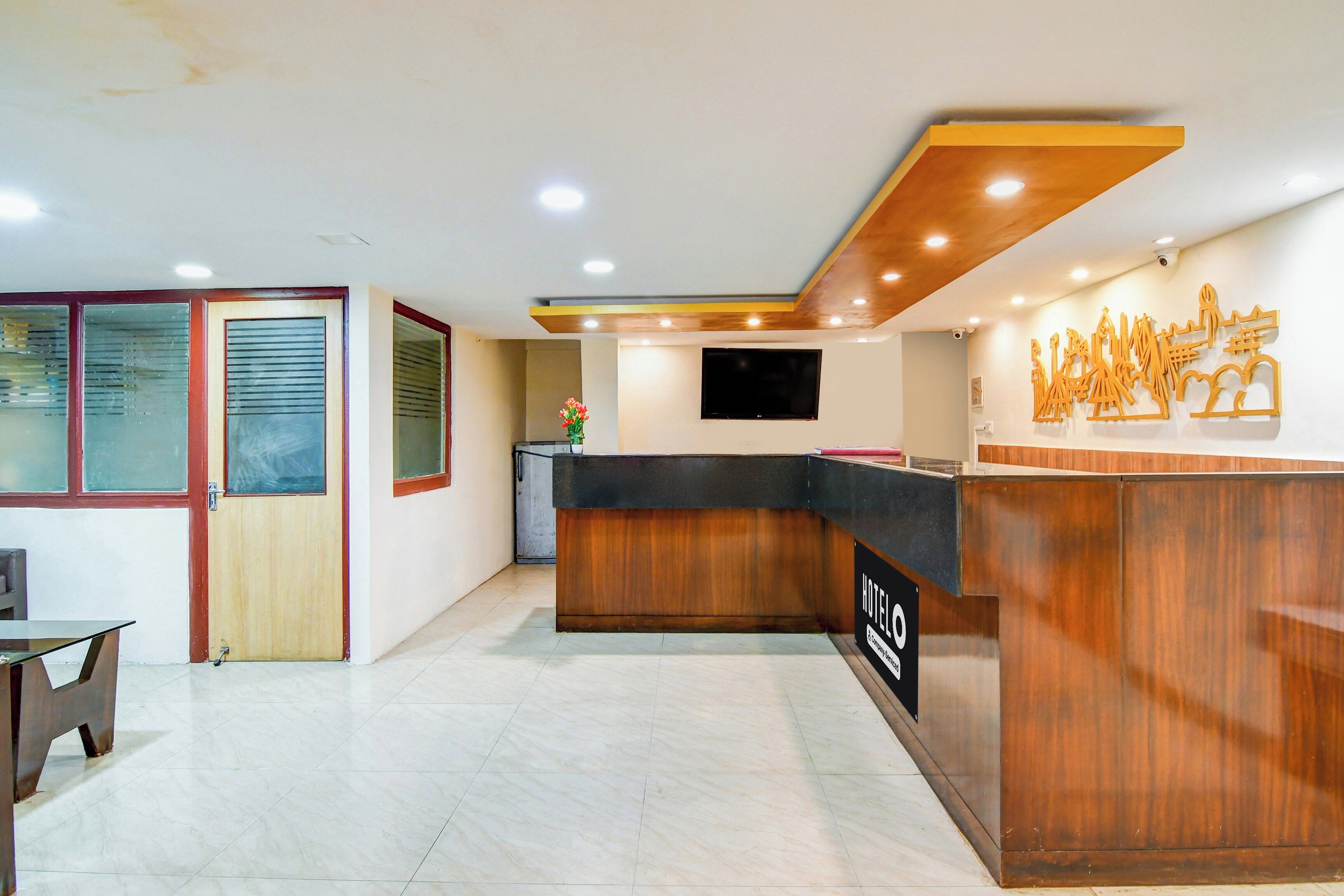 A hotel reception area with a wooden reception desk, a flat-screen TV mounted on the wall, and a decorative wall piece.
