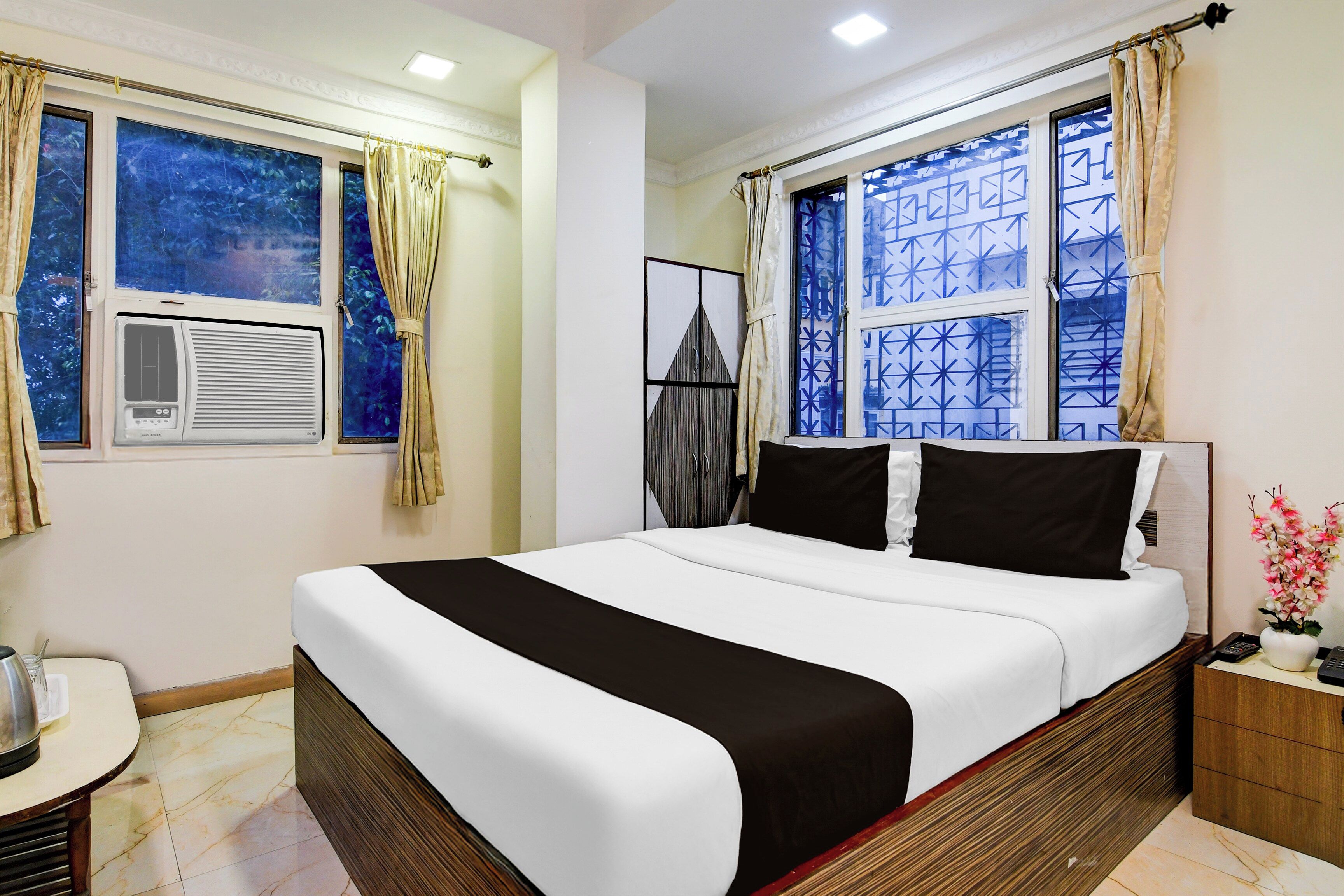 A hotel room with a bed, a nightstand, a window with curtains, and a wall-mounted air conditioner.