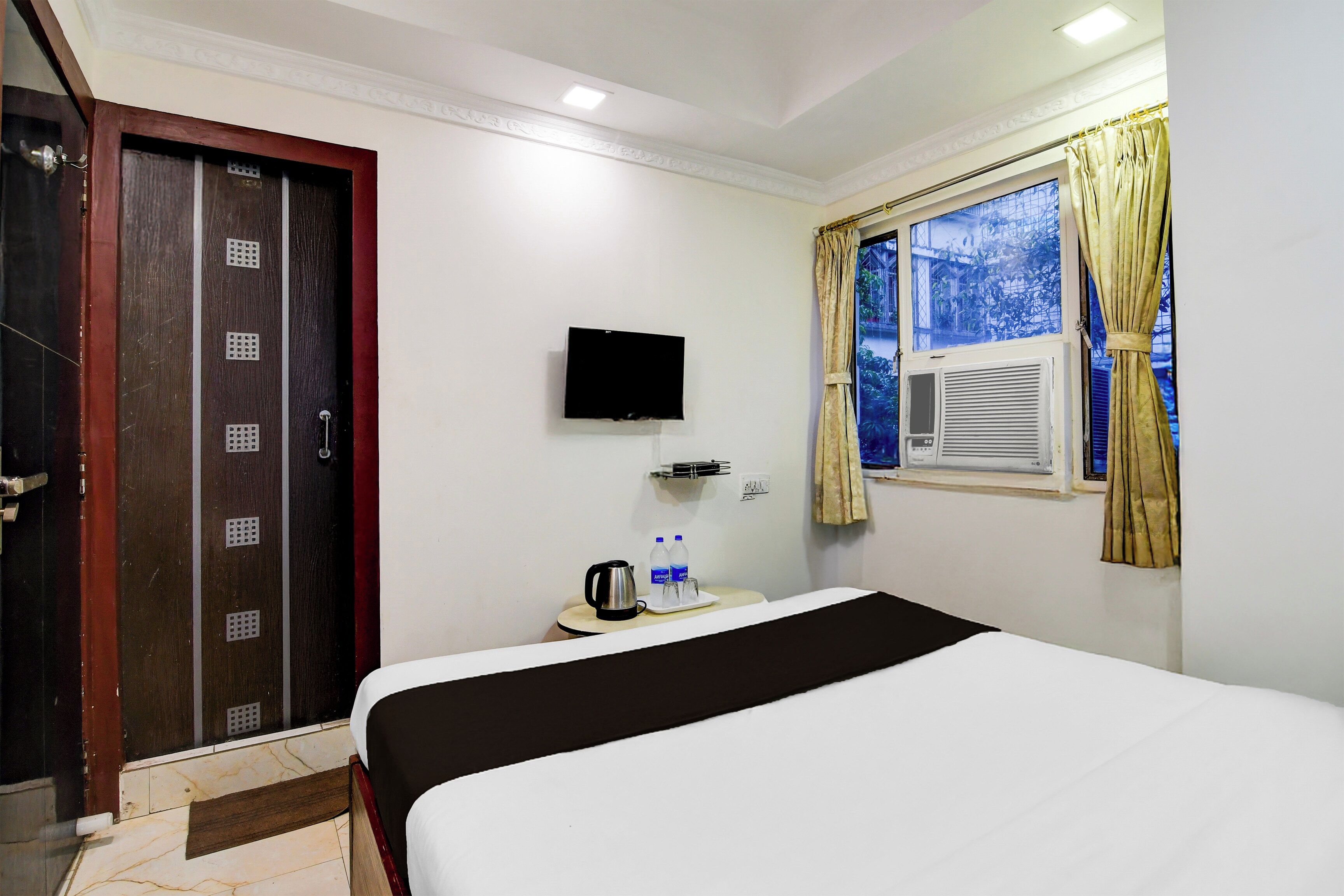 A hotel room with a bed, a television mounted on the wall, a window with curtains, and a door with a security lock.