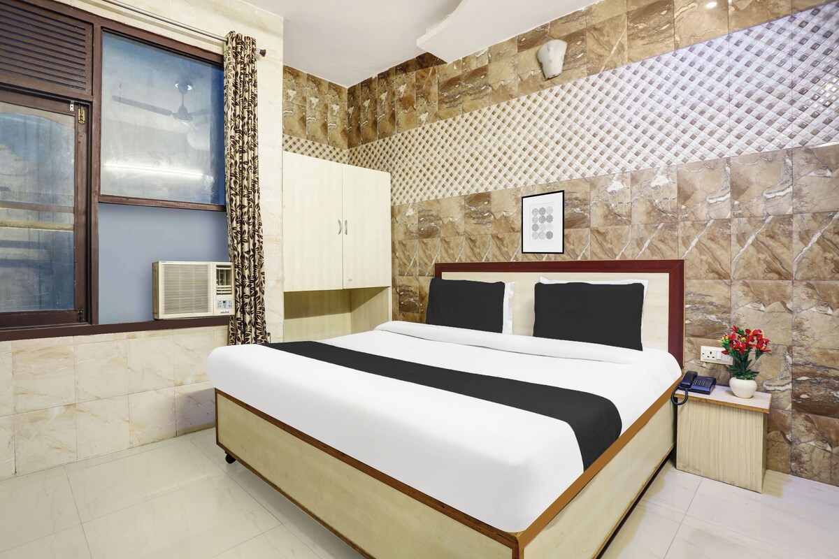 A hotel room with a bed, a wardrobe, a window with curtains, a wall-mounted air conditioner, and a small bedside table with a phone and a vase of flowers.