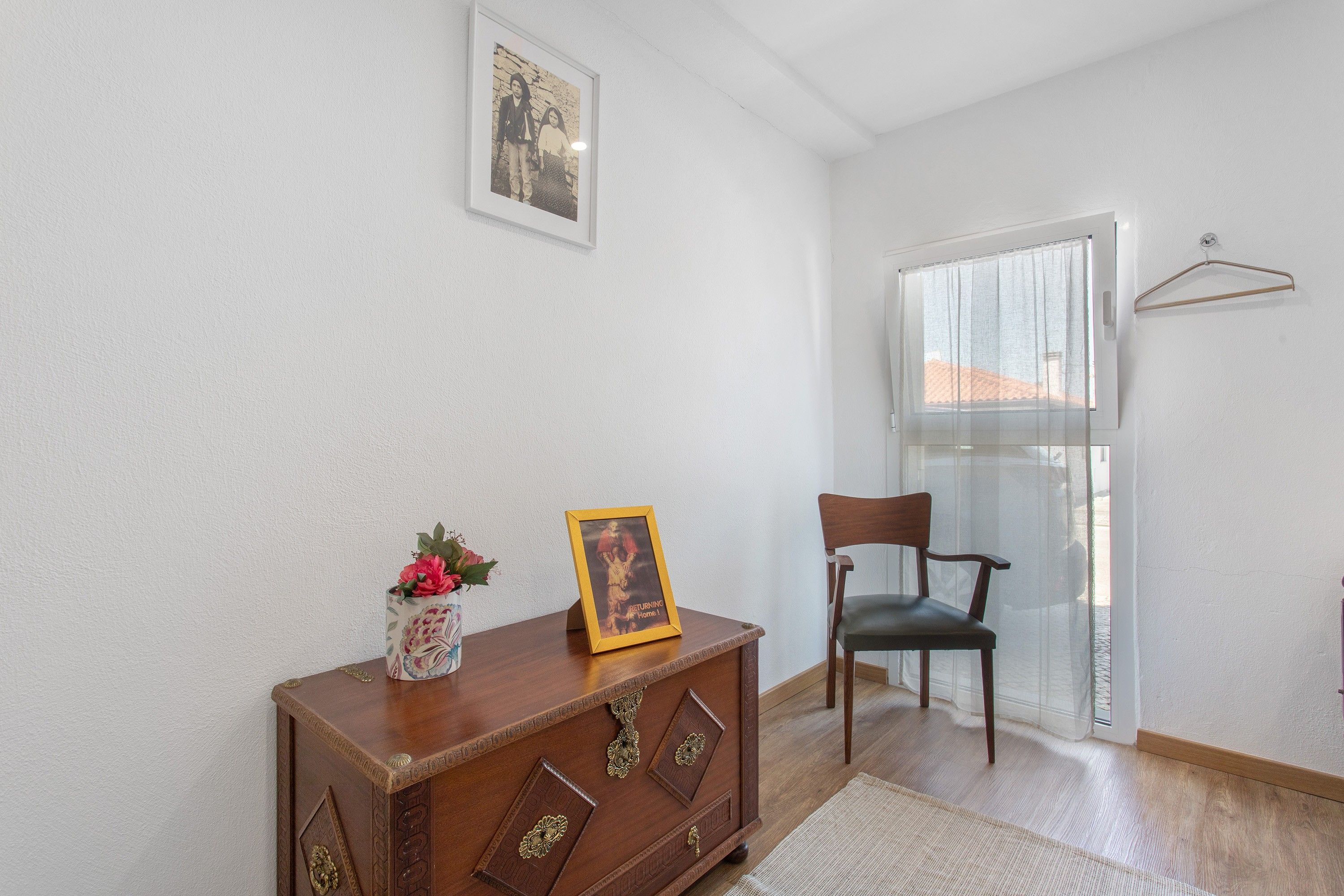 Una habitación con un armario de madera, una silla, un cuadro enmarcado y una ventana con cortina.