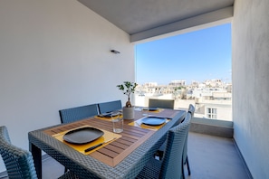 Ein Balkon mit Esstisch für zwei Personen, Korbstühlen, einem Blumentopf und Blick auf die Stadt.