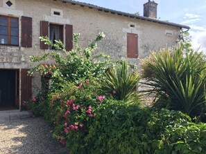 Un bâtiment rustique en pierre, doté de volets en bois, d’une allée en gravier et d’une végétation luxuriante comprenant des plantes à fleurs et un grand agave.