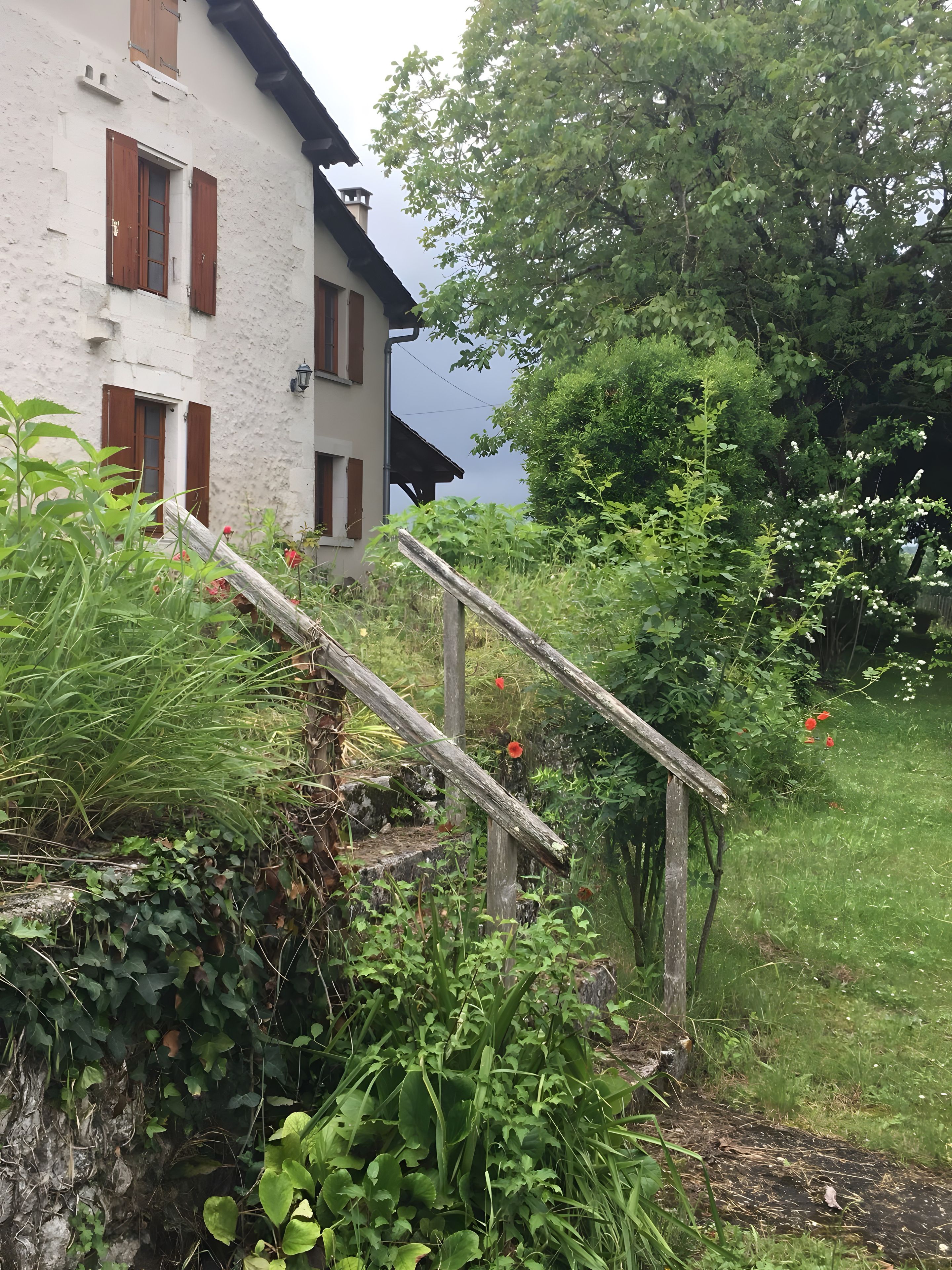 Un bâtiment rustique avec une façade en pierre et des volets en bois, entouré d’une végétation luxuriante et agrémenté d’un escalier en bois menant à un niveau supérieur.
