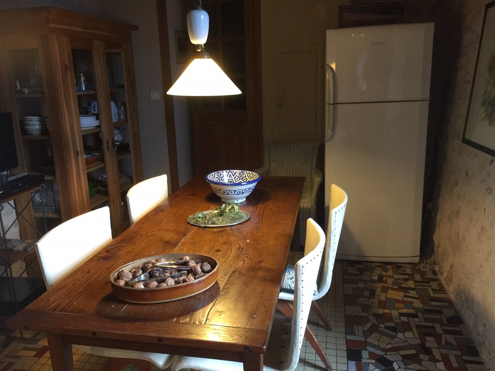 Une table de salle à manger en bois, avec des chaises, un bol de noix et un bol décoratif. Un réfrigérateur se trouve à l’arrière-plan.