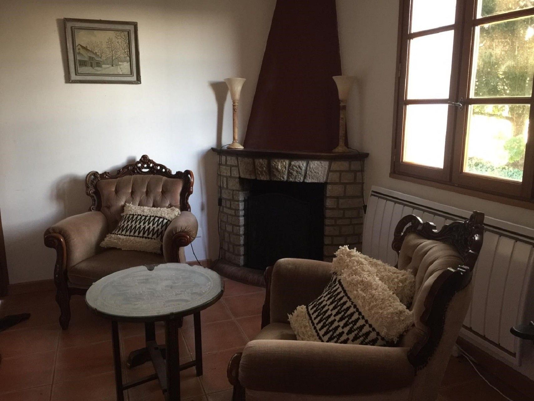 A cozy living room with a fireplace, two armchairs, a small round table, and a window with natural light.