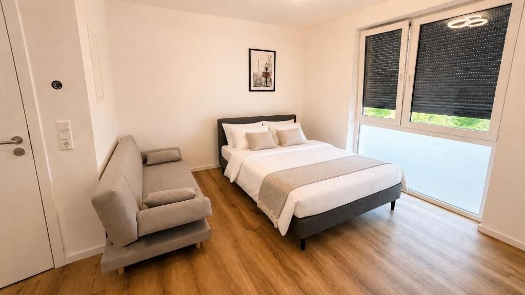 A bedroom with a bed, a sofa, a window with blinds, and a door.