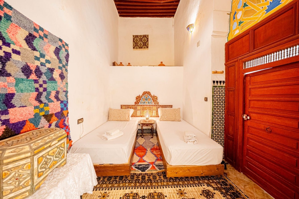 A small, cozy room with two beds, a patterned rug, a wooden cabinet, and a red door.