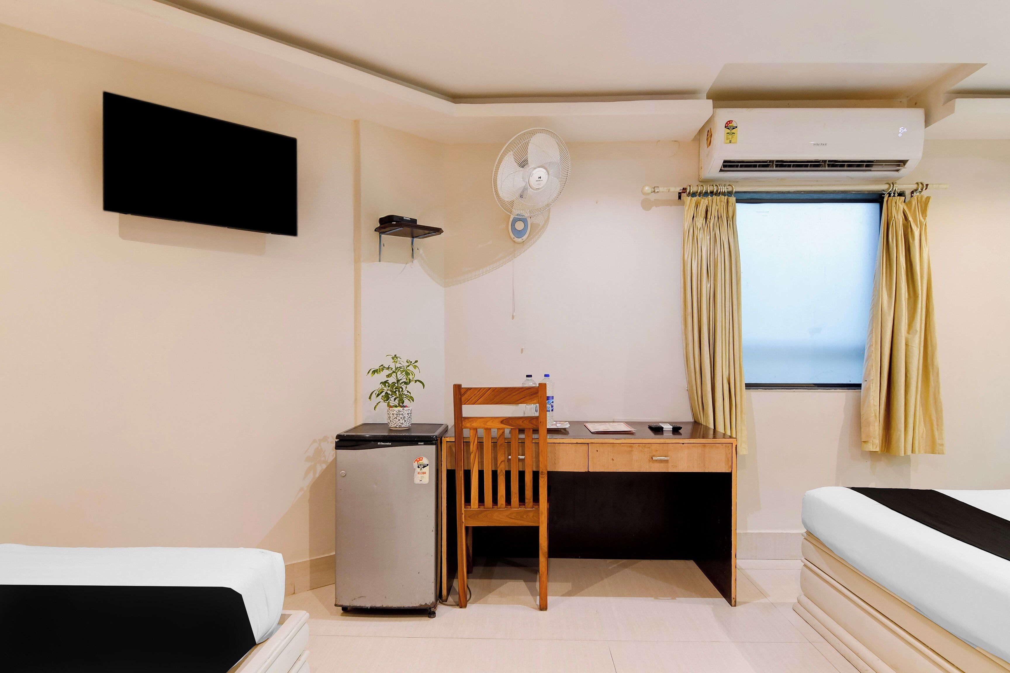 A compact room with a bed, a desk, a chair, a small refrigerator, a wall-mounted fan, and an air conditioning unit.