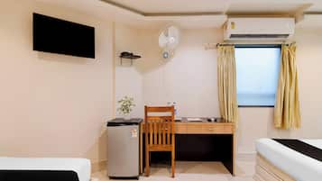Quarto compacto com cama, escrivaninha, cadeira, pequeno frigorífico, ventoinha fixada na parede e ar condicionado.