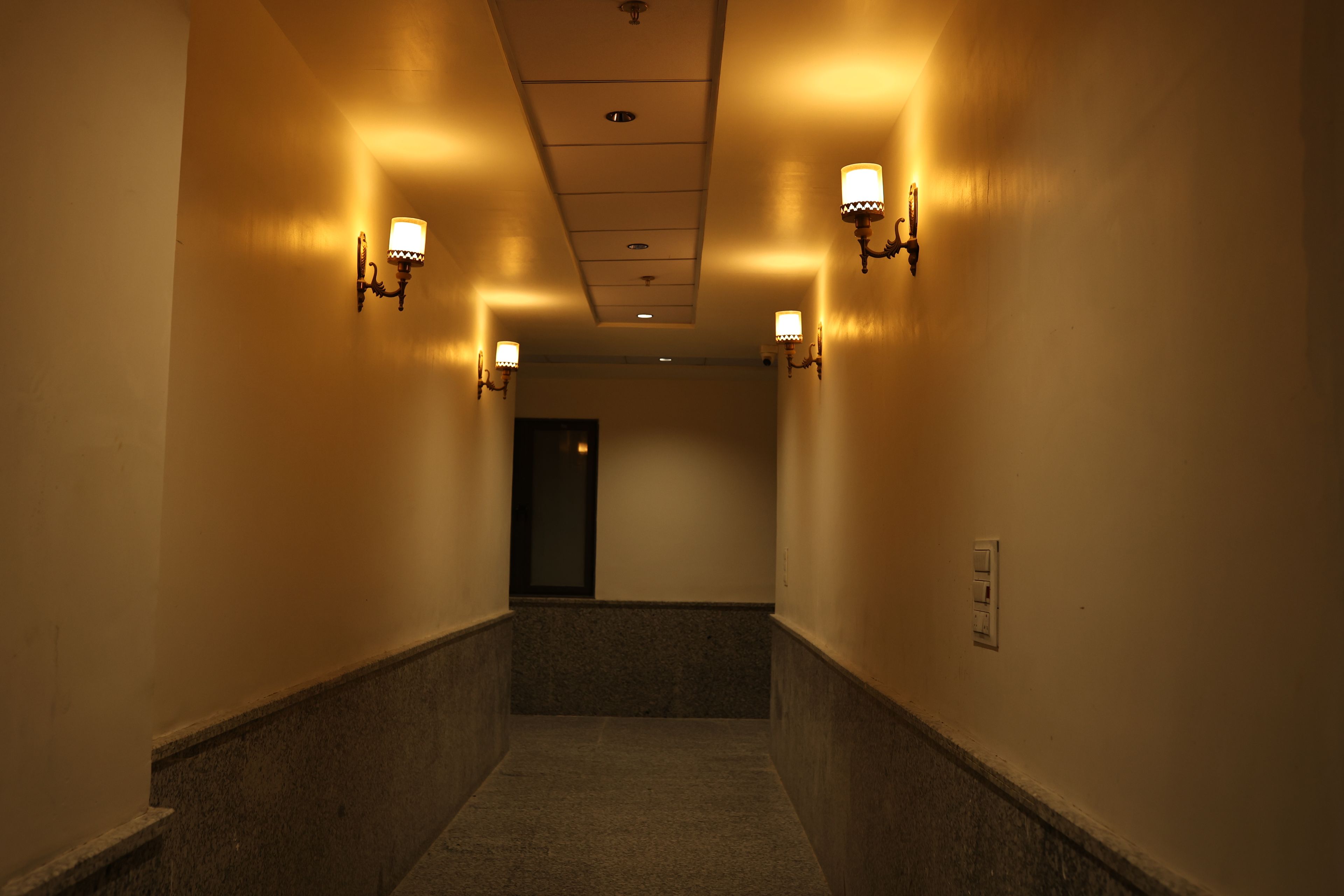 A corridor with wall-mounted sconces, a ceiling light, and a door at the end.